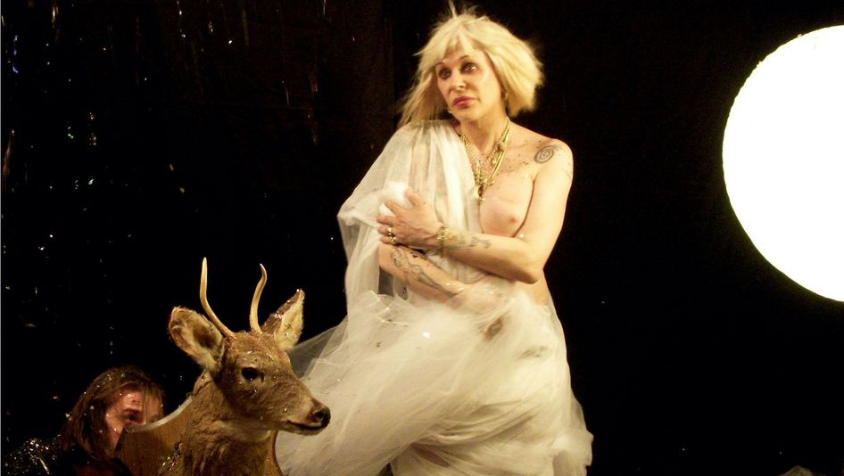 Film still from THE BALLAD OF GENESIS AND LADY JAYE: Genesis Breyer P-Orridge stands in front of a black background with a few points of light, dressed in a tulle cape, one breast exposed.