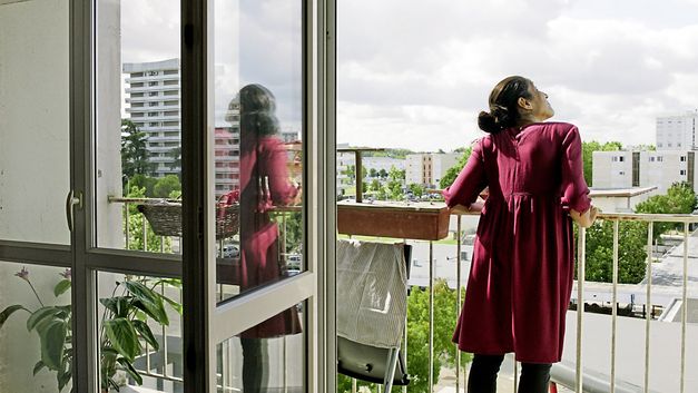 Still from the film "Europe" by Philipp Scheffner. A woman in a red dress is leaning against the balcony looking up.  