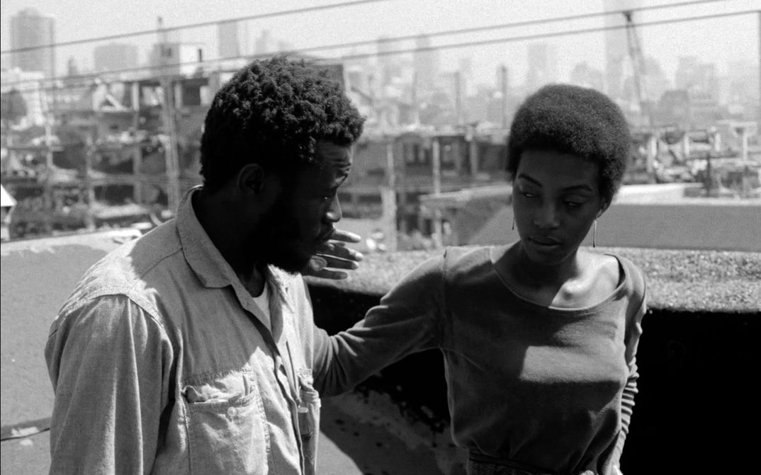 Filmstill aus BUSHMAN: Ein Mann und eine Frau gehen eine Straße entlang, sie streicht ihm über die Wange.