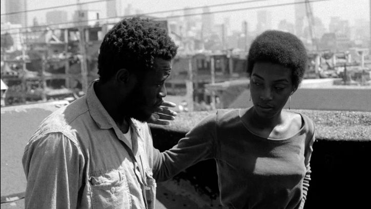 Film still from BUSHMAN: A man and a woman walk along a street, she strokes his cheek.