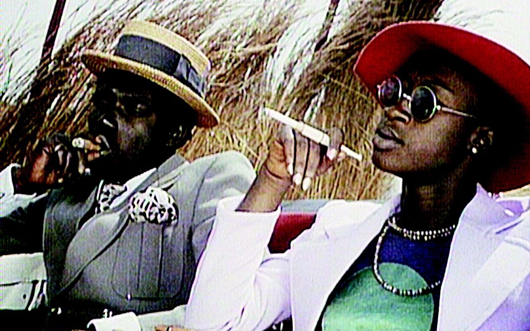 Filmstill aus TOUKI BOUKI: Zwei glamourös gekleidete Menschen sitzen rauchend in der Sonne.