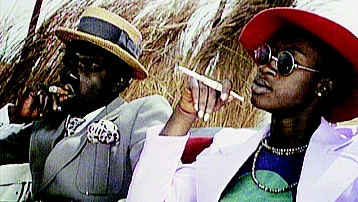 Filmstill aus TOUKI BOUKI: Zwei glamourös gekleidete Menschen sitzen rauchend in der Sonne.