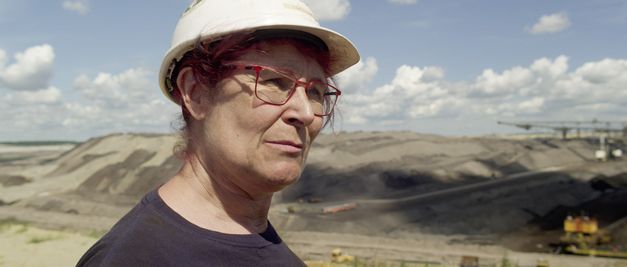 Film still from STOLZ & EIGENSINN by Gerd Kroske. It shows a woman with a safety helmet and red glasses in front of an open-cast mine.