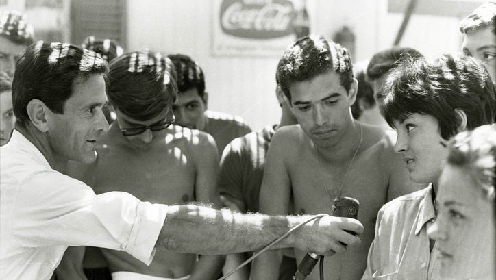 Filmstill aus COMIZI D´AMORE: Eine Gruppe von Menschen. Eine Mann hält ein Mikrofon zu einer Person.
