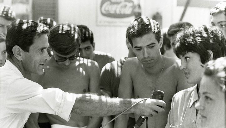 Filmstill aus COMIZI D´AMORE: Eine Gruppe von Menschen. Eine Mann hält ein Mikrofon zu einer Person.