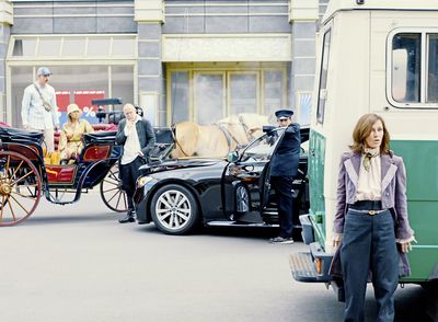 Filmstill aus „L’état et moi“ von Max Linz. Eine Frau versteckt sich hinter einem Polizeitransporter. Dahinter sehen wir eine Touristenpärchen in einer Kutsche und den uniformierten Fahrer einer schwarzen Limousine. Sie schauen in ihre Richtung. 