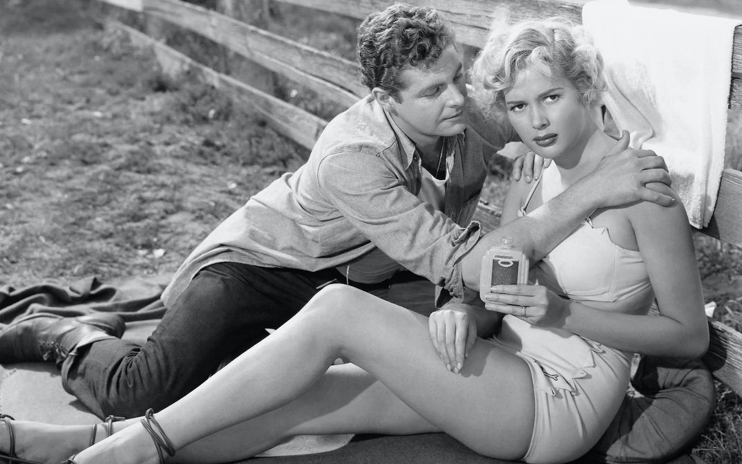 Filmstill aus PICKUCK: Eine junge, leichtbekleidete Frau sitzt an einen Zaun gelehnt. Ein Mann sitzt neben ihr und greift sie an die Schultern.