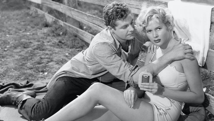 Film still from PICKUCK: A young, scantily clad woman sits leaning against a fence. A man sits next to her and grabs her by the shoulders.