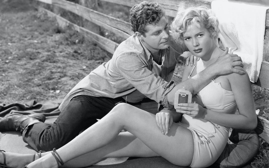 Filmstill aus PICKUCK: Eine junge, leichtbekleidete Frau sitzt an einen Zaun gelehnt. Ein Mann sitzt neben ihr und greift sie an die Schultern.
