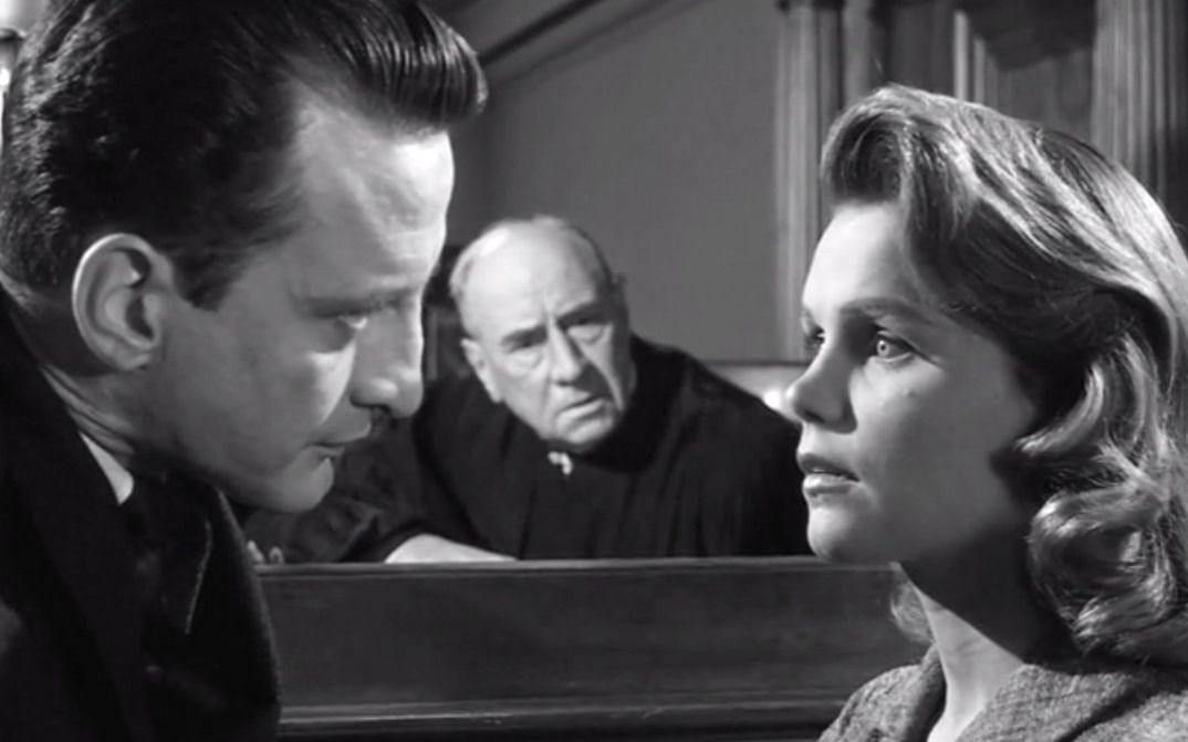 Filmstill from ANATOMY OF A MURDER: A man and a woman look at each other. The woman looks startled. Another man is sitting in the background looking at the two of them.