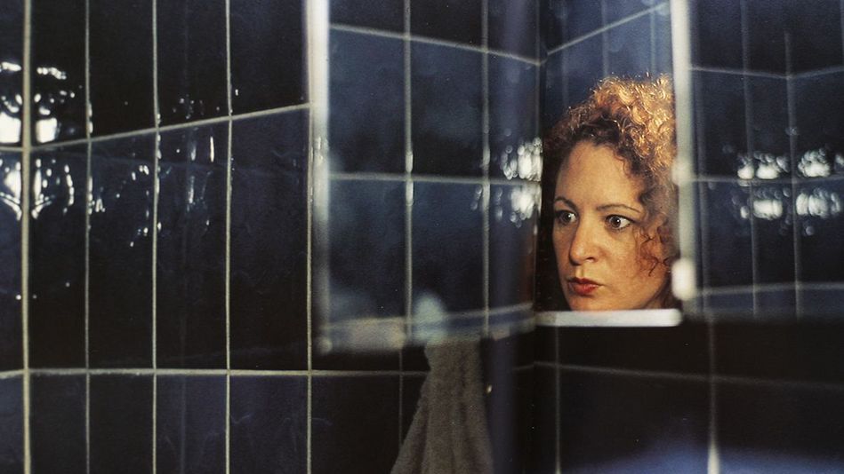 Filmstill aus I'LL BE YOUR MIRROR: In einem dunkelblau gekachelten Badezimmer ist das Gesicht von Nan Goldin in einem kleinen Spiegel zu sehen.