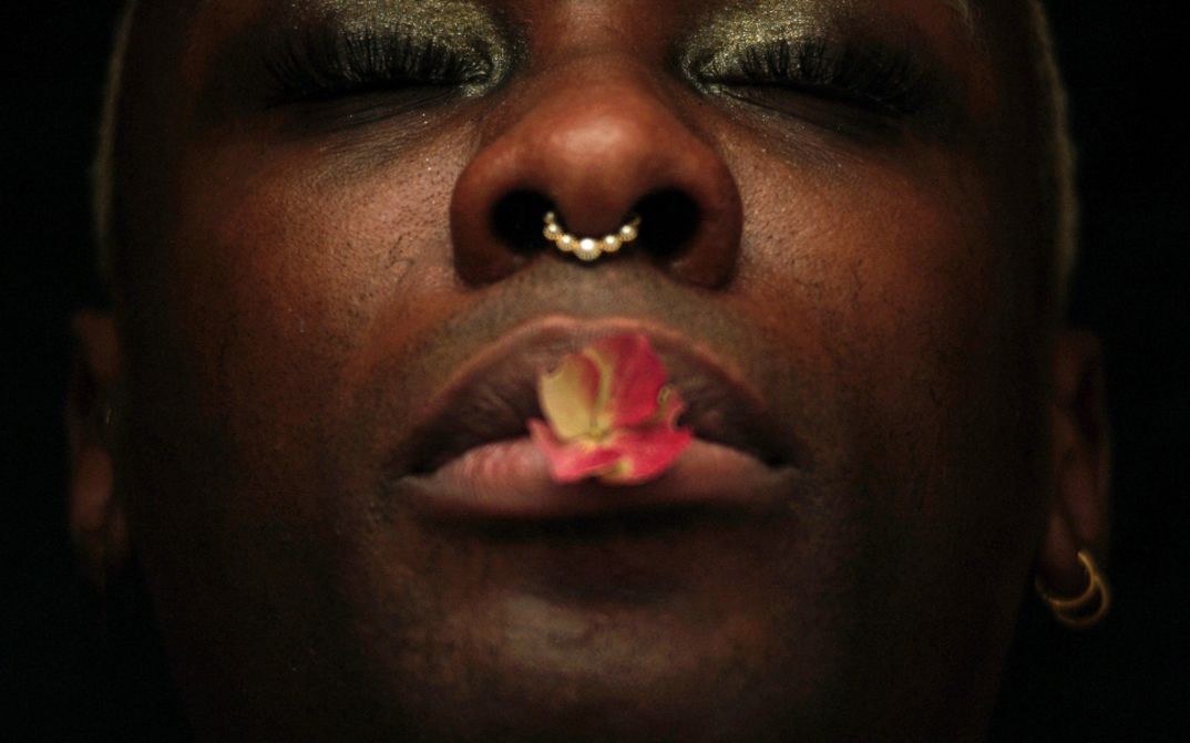 Extreme Nahaufnahme eines Gesichts mit geschlossenem Mund, Nasenpiercing und einer Blüte auf den Lippen.