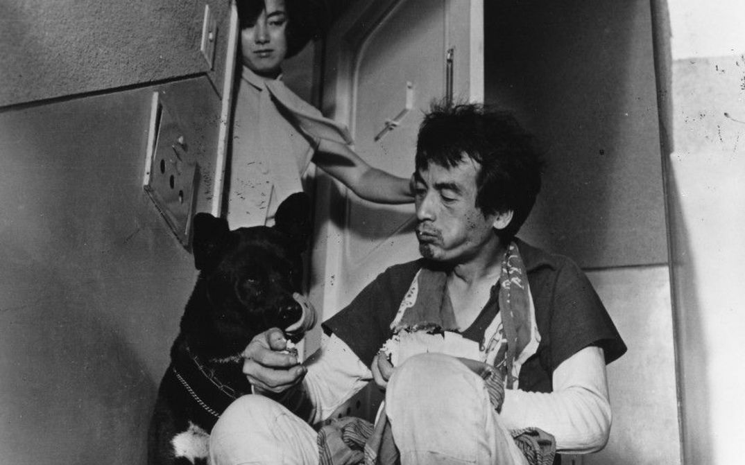 A man is sitting next to a dog on a landing. A woman looks on from the open front door.Film still from SHE AND HE: