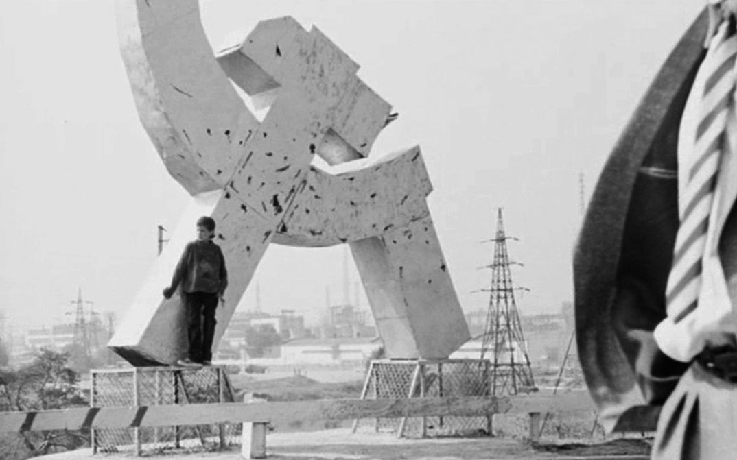Filmstill aus SCHWANENSEE. ZONE: Eine Person lehnt an ein Hammer-und-Sichel-Monument.