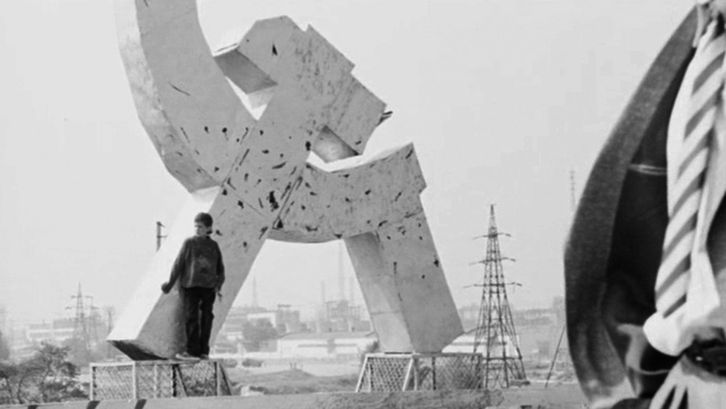 Film still from SWAN LAKE. THE ZONE: A person leans against a hammer and sickle monument.