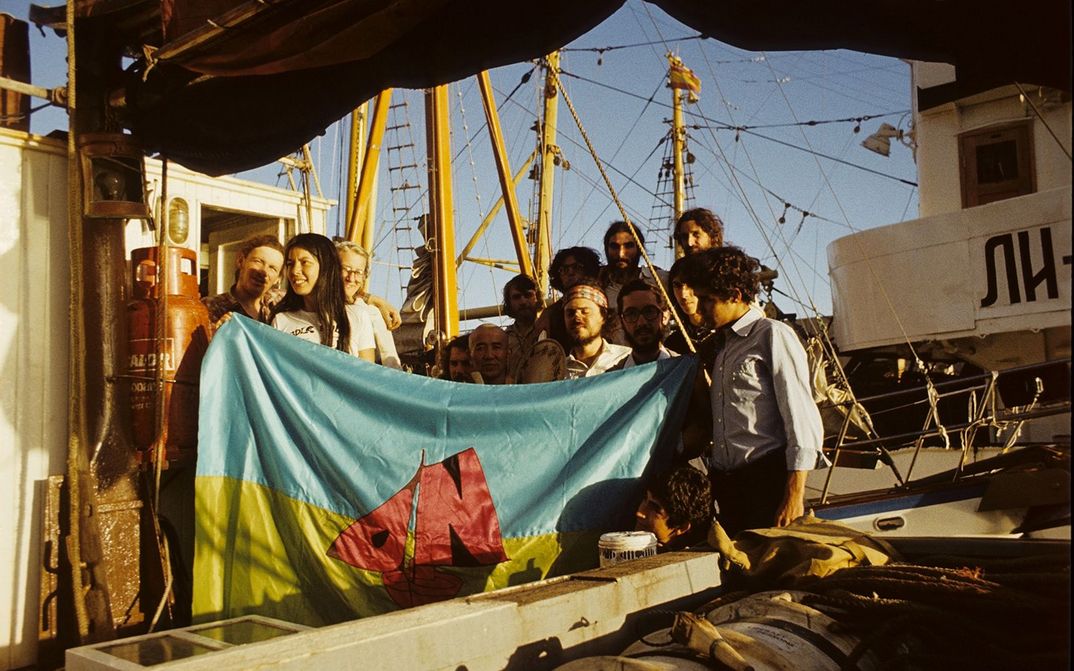 Filmstill from OPERATION NAMIBIA:  A group of people are standing on a sailing ship. They look happy and are holding a flag in blue and yellow and a red sailing ship.