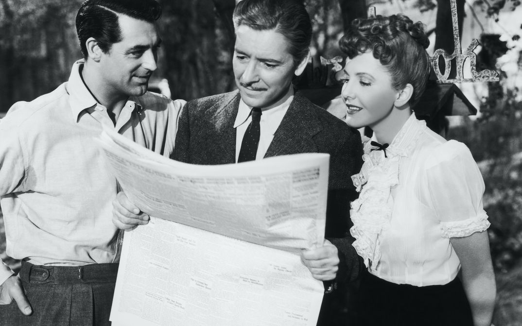 Filmstill aus THE TALK OF THE TOWN: Zwei Männer und eine Frau stehen zusammen und blicken auf eine Zeitung.