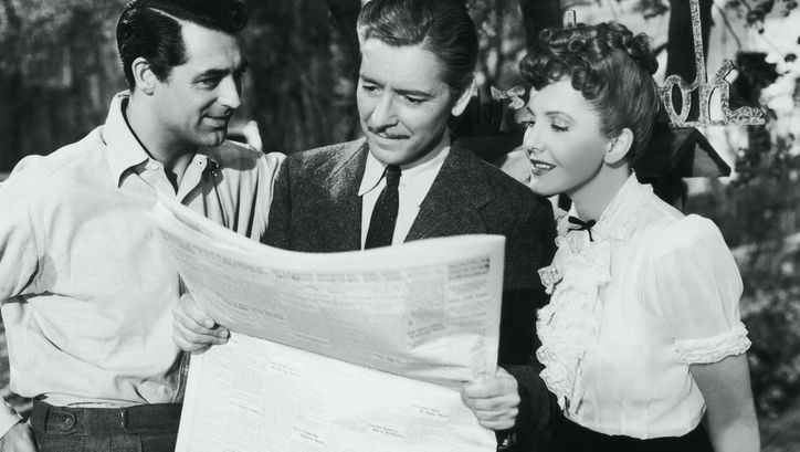 Filmstill aus THE TALK OF THE TOWN: Zwei Männer und eine Frau stehen zusammen und blicken auf eine Zeitung.