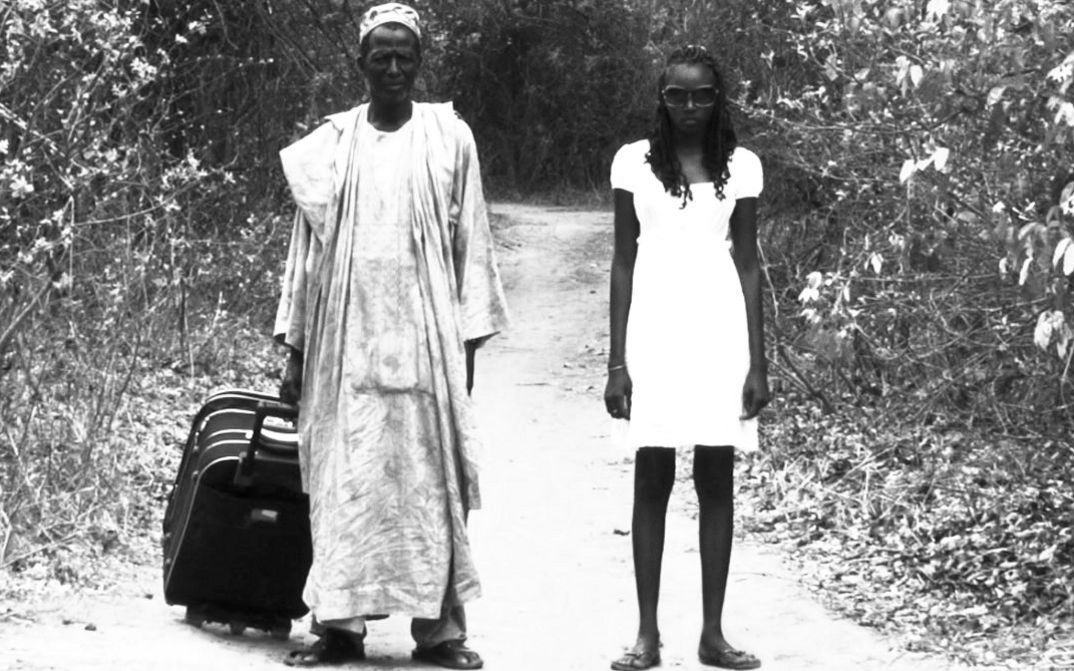 Film still from A BATALHA DE TABATÔ: A man and a younger woman stand next to each other on a country lane and look towards the camera. He is pulling a suitcase behind him.