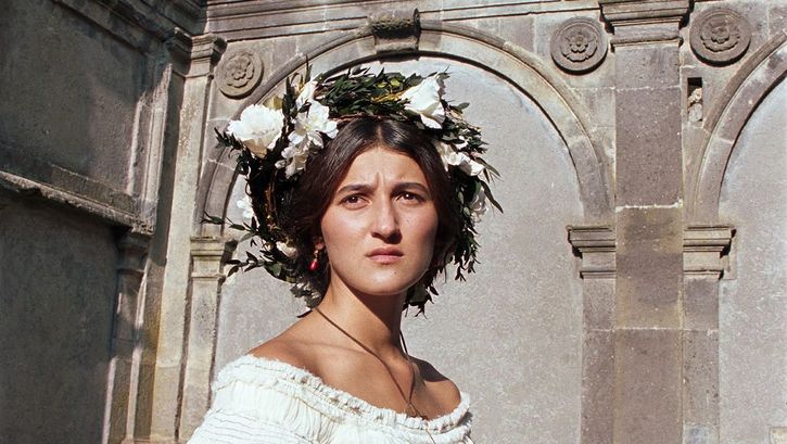 Film still from RE GRANCHIO. A woman in a white dress and flowers in her hair looks into the distance, frowning.