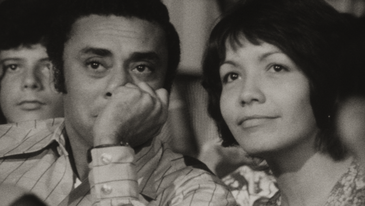 Film still from DE CIERTA MANERA: A woman and a man sit side by side and look at a screen off-screen.