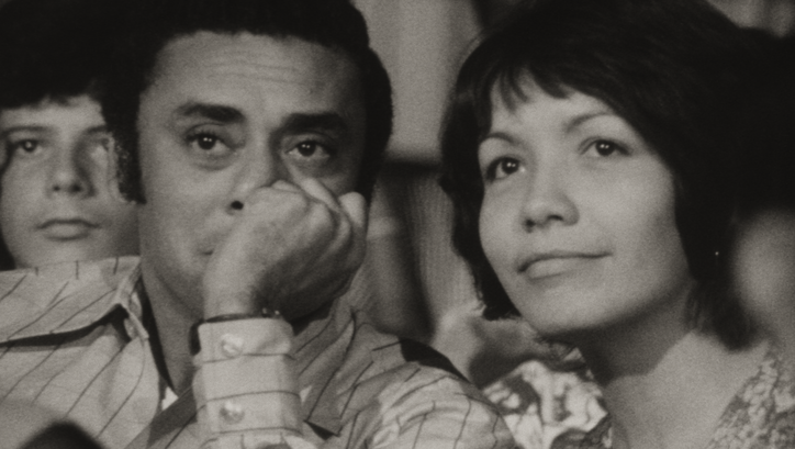 Film still from DE CIERTA MANERA: A woman and a man sit side by side and look at a screen off-screen.