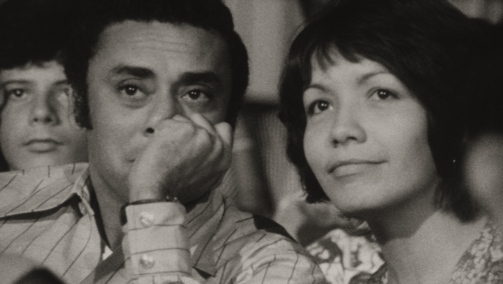 Film still from DE CIERTA MANERA: A woman and a man sit side by side and look at a screen off-screen.