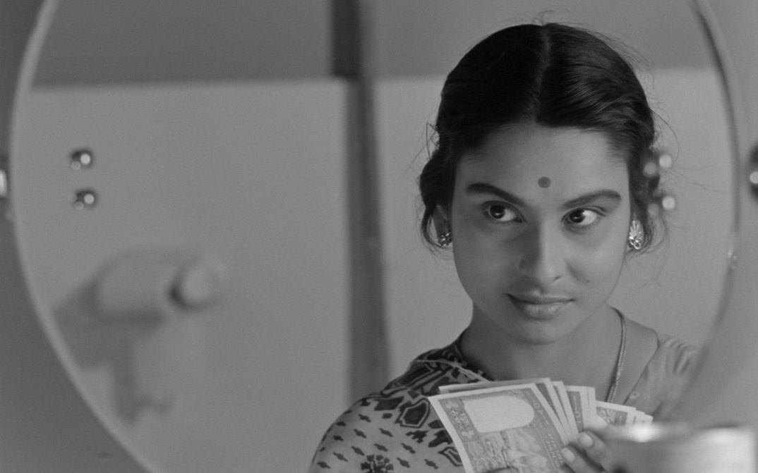 Filmstill from MAHANAGAR: A woman with a dot on her forehead looks in the mirror and holds a stack of banknotes in her hands.