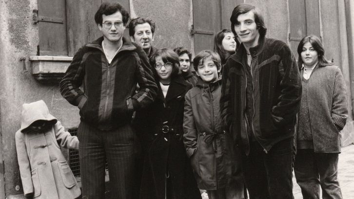 Film still from FAMILIE VILLANO KEHRT NICHT ZURÜCK: Several adults and children are standing on the street in front of a house wall.