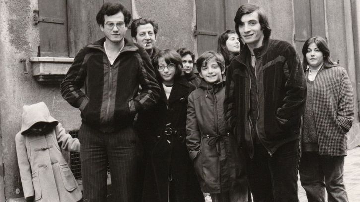 Filmstill aus FAMILIE VILLANO KEHRT NICHT ZURÜCK: Mehrere Erwachsene und Kinder stehen auf der Straße vor einer Hausmauer.