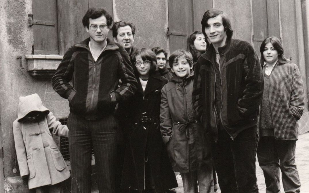 Filmstill aus FAMILIE VILLANO KEHRT NICHT ZURÜCK: Mehrere Erwachsene und Kinder stehen auf der Straße vor einer Hausmauer.
