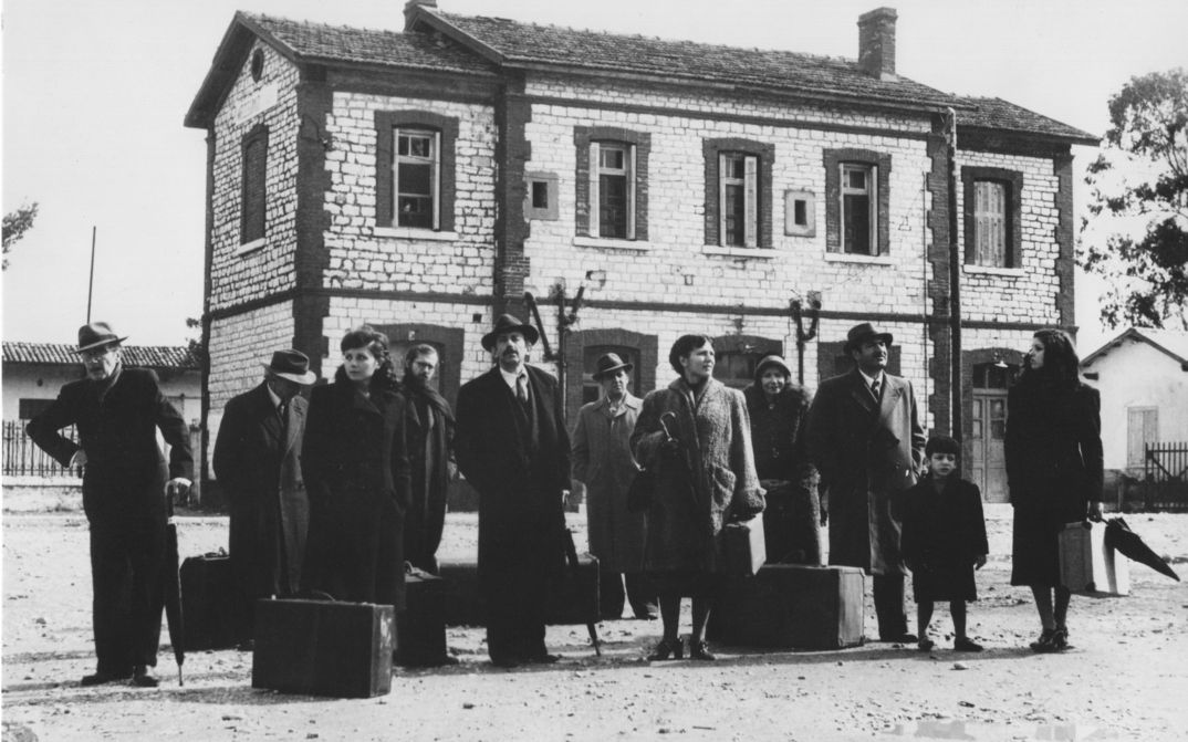 Filmstill aus O THIASSOS: Eine Gruppe von Menschen mit Koffern steht vor einem Gebäude.