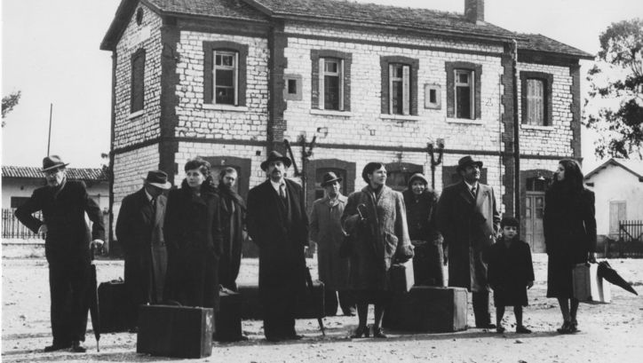 Film still from O THIASSOS: A group of people with suitcases stands in front of a building.