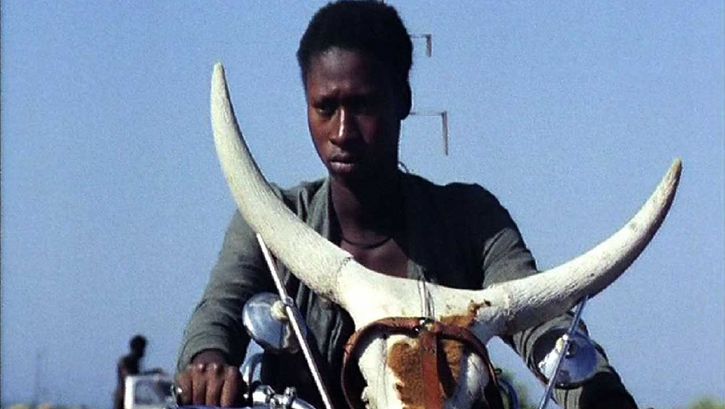 Filmstill aus TOUKI BOUKI: Eine junge Person auf einem Motorrad, auf dem ein Kuhschädel befestigt ist.