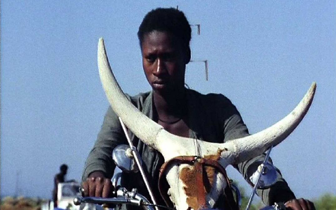 Filmstill aus TOUKI BOUKI: Eine junge Person auf einem Motorrad, auf dem ein Kuhschädel befestigt ist.