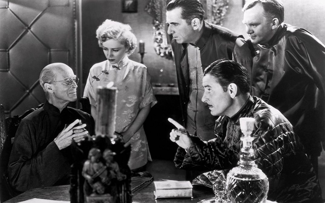 Film still from LOST HORIZON: A group of people are sitting and standing around a table. Everyone is looking at one person.