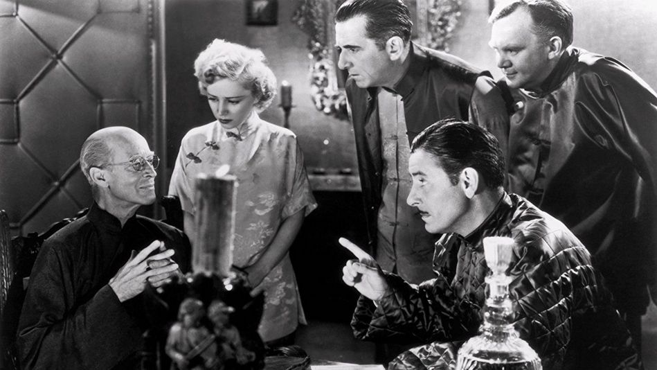 Film still from LOST HORIZON: A group of people are sitting and standing around a table. Everyone is looking at one person.