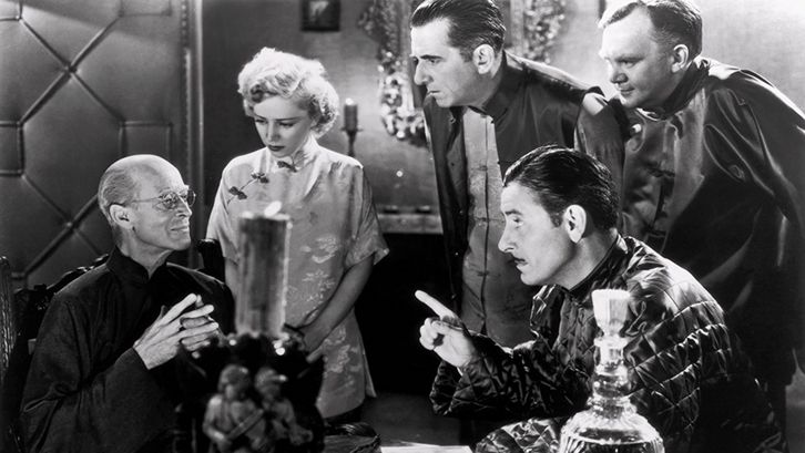 Film still from LOST HORIZON: A group of people are sitting and standing around a table. Everyone is looking at one person.