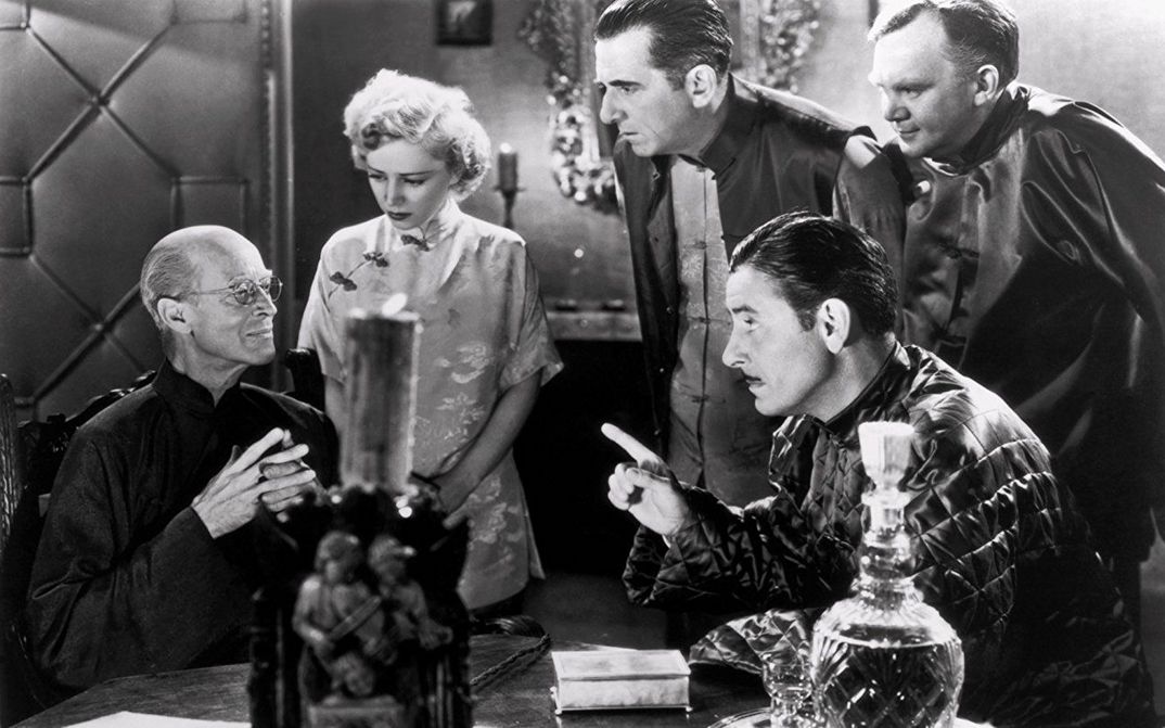 Filmstill aus LOST HORIZON: Eine Gruppe von Menschen sitzt und steht um einen Tisch herum. Alle blicken auf eine Person.