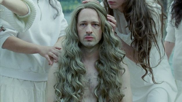 Filmstill aus „Mammalia“ von Sebastian Mihăilescu. Eine Nahaufnahme eines Mannes mit Bart und langem Haar, der in die Kamera schaut. Um ihn herum stehen Menschen, die ihn berühren.