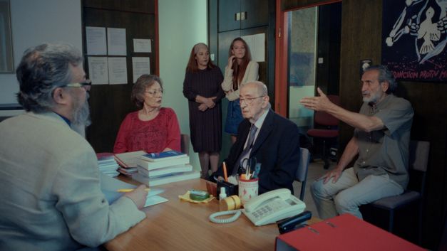 Film still from RESTITUCIJA, ILI, SAN I JAVA STARE Gare by Želimir Žilnik. It shows an office space with six people. Four people are sitting in the front. Two women stand in the background and observe the situation.