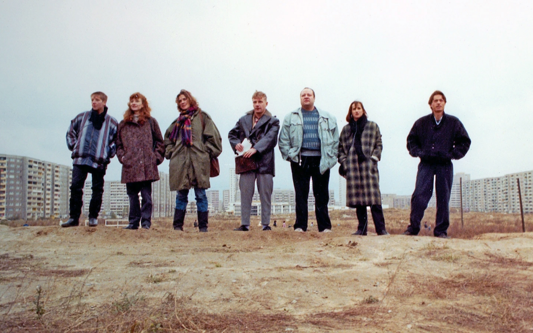 Film still from DIE ARCHITEKTEN: Seven people stand next to each other on a mound of earth and look ahead. Some buildings can be seen in the background.