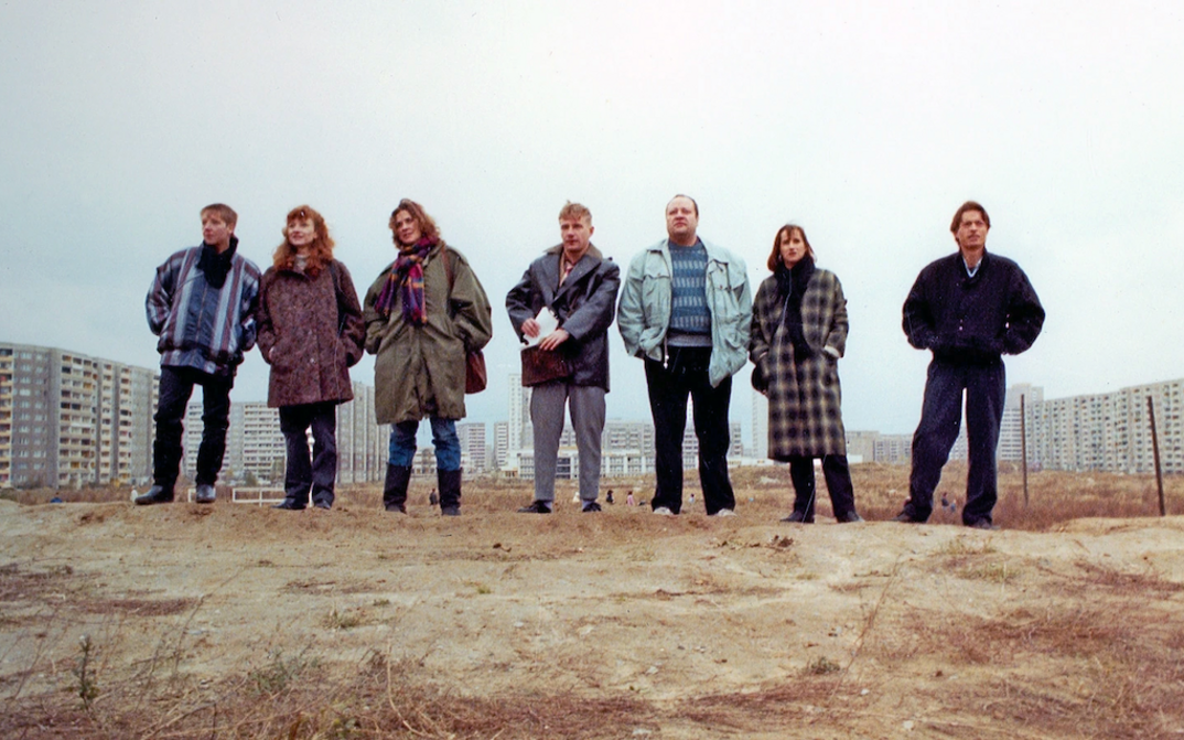 Film still from DIE ARCHITEKTEN: Seven people stand next to each other on a mound of earth and look ahead. Some buildings can be seen in the background.