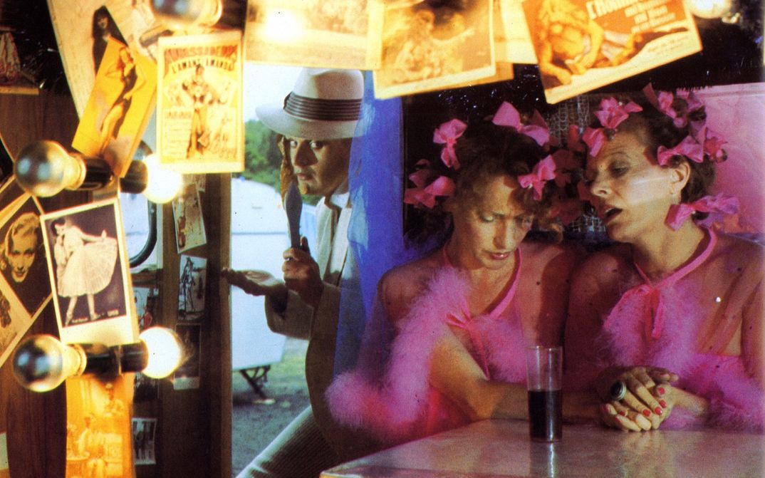 Film still from FREAK ORLANDO: Two women wearing identical pink dresses and pink bows in their hair are sitting in a bar lavishly decorated with photos and lamps.