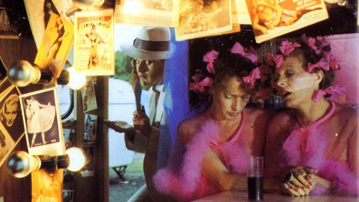 Film still from FREAK ORLANDO: Two women wearing identical pink dresses and pink bows in their hair are sitting in a bar lavishly decorated with photos and lamps.