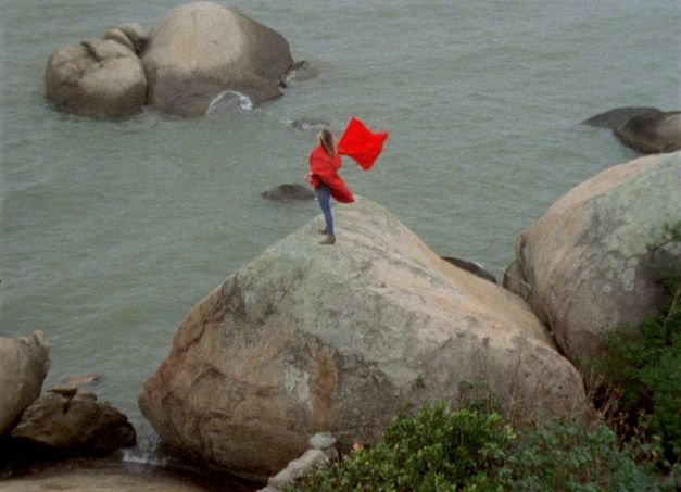 Filmstill aus ORÁCULO: Eine Person mit rotem Mantel und roter Fahne steht auf einem Felsen am Meer.