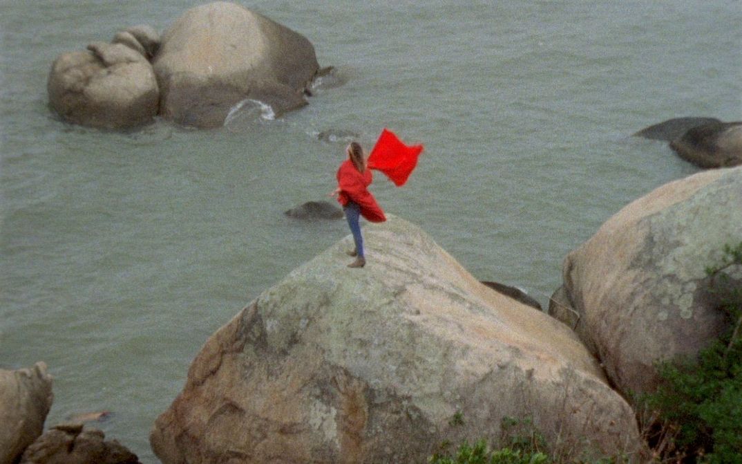 Film still from ORÁCULO: A person with a red coat and a red flag is standing on a rock by the sea.