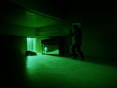 Still from the film "O trio em mi bemol (The Kegelstatt Trio)" by Rita Azevedo Gomes. A man with a crowbar approaches a glowing green light in a dark room. 