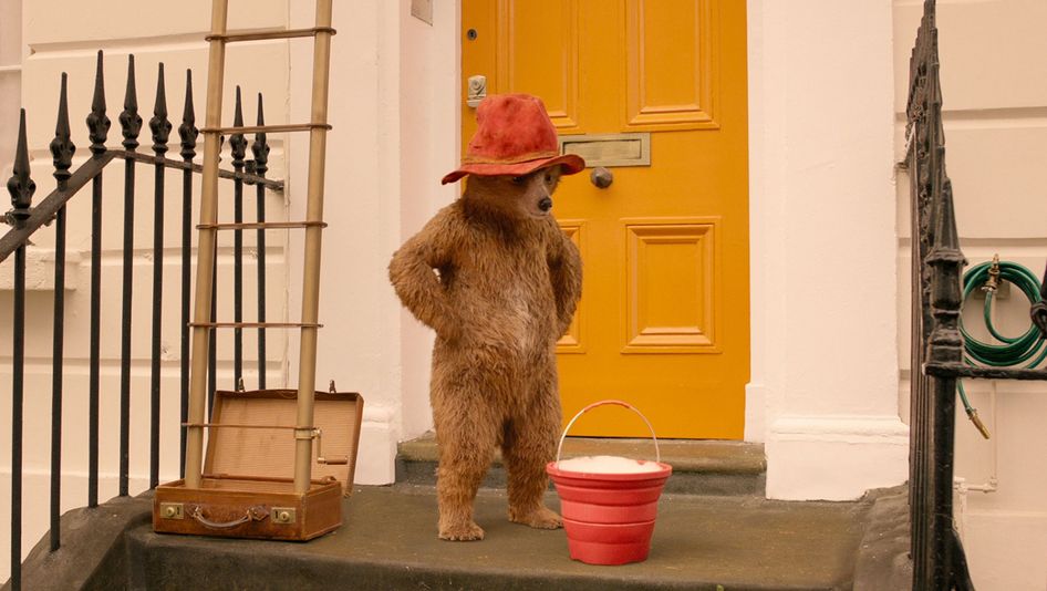 Filmstill aus PADDINGTON 2: Der Teddybär Paddington trägt einen roten Hut und steht vor der gelben Tür eines Hauses.