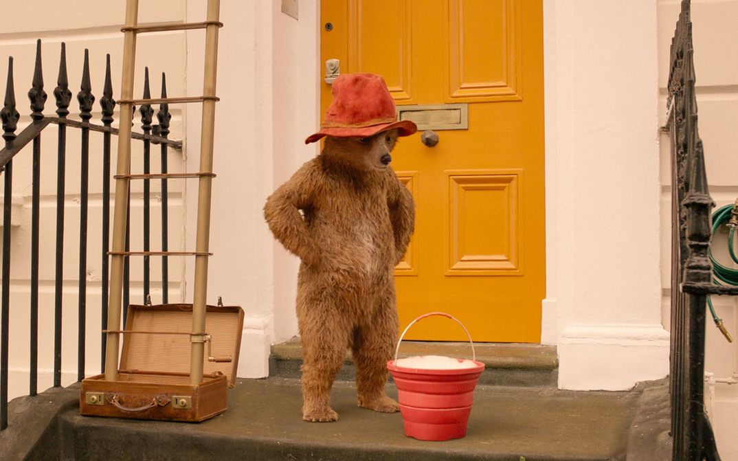 Filmstill aus PADDINGTON 2: Der Teddybär Paddington trägt einen roten Hut und steht vor der gelben Tür eines Hauses.